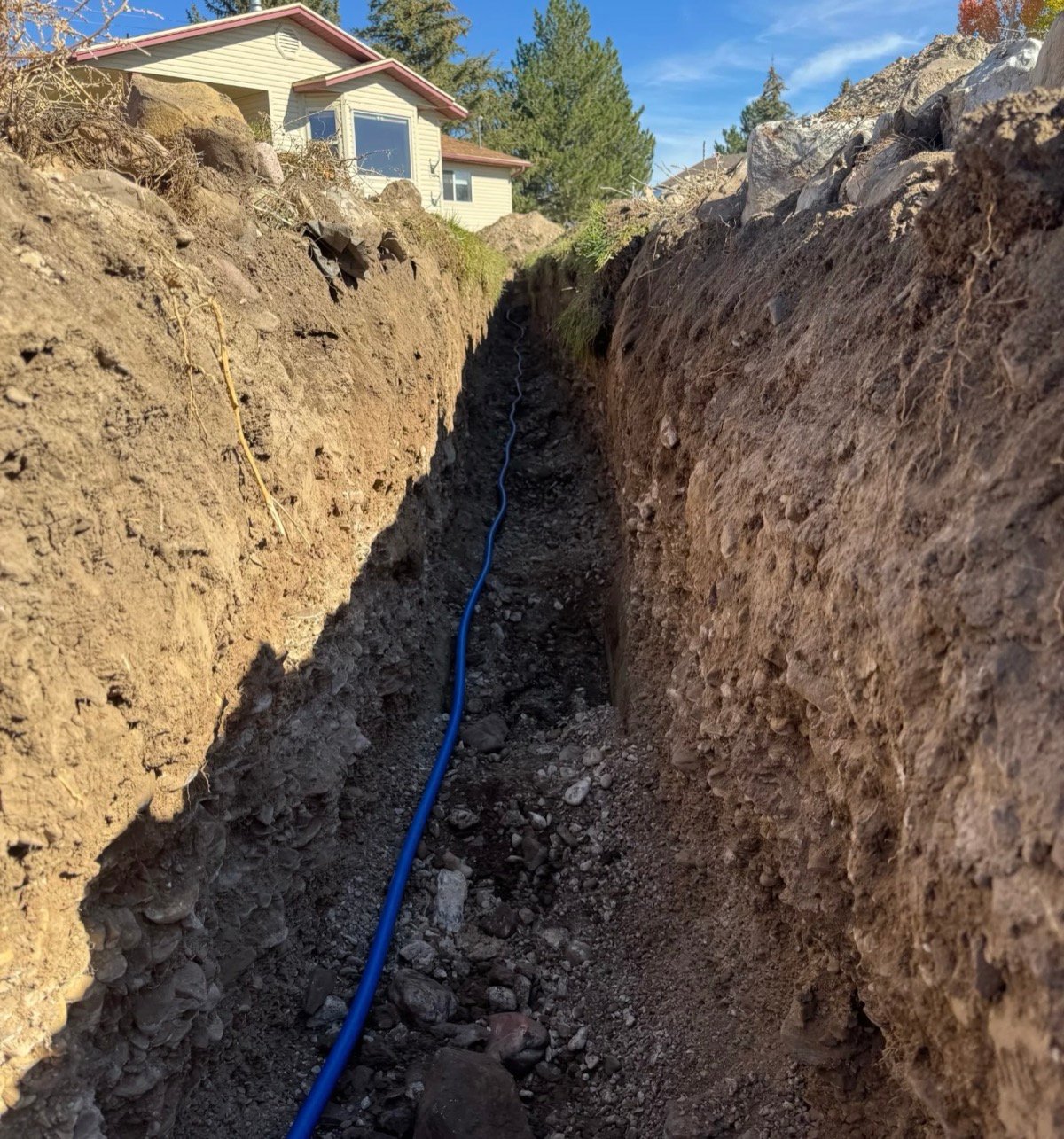 Deep open trench excavation for water line replacement showing new blue PEX pipe in trench